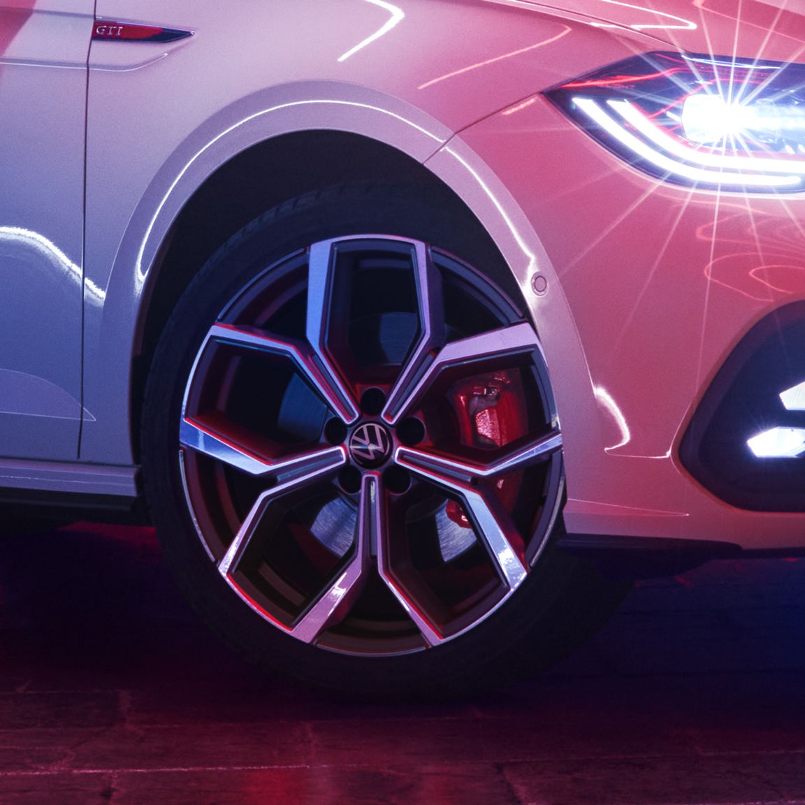 Detailný záber pravého predného kolesa bieleho VW Polo GTI. Koleso má výrazný moderný dizajn. Svetlá auta sú zapnuté a v lakovaní vozidla sa odráža okolité svetlo.