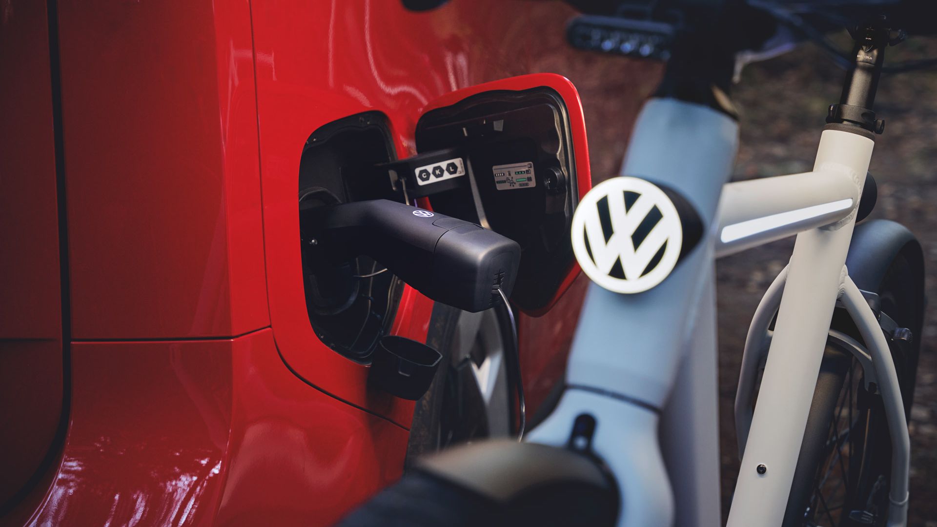 Záber zblízka na červené elektrické auto, ktoré je prostredníctvom nabíjacieho kábla pripojené k nabíjacej zásuvke. V popredí stojí biely elektrobicykel s jasne viditeľným logom VW na riadidlách. Scéna vyjadruje udržateľnú mobilitu pomocou auta a bicykla.