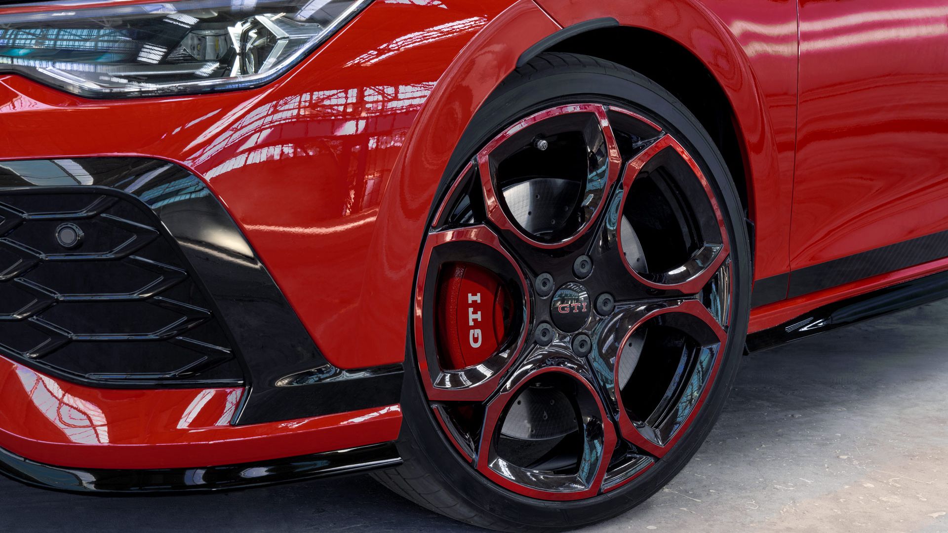 Detailný záber červeného modelu Golf GTI s výrazným čiernočerveným diskom s logom GTI. Predný nárazník je čiastočne viditeľný. Auto je zaparkované v jasnom prostredí s dostatkom svetla.