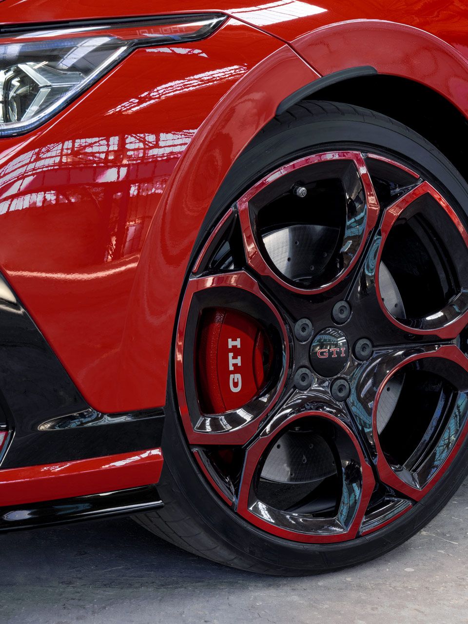 Detailný záber červeného modelu Golf GTI s výrazným čiernočerveným diskom s logom GTI. Predný nárazník je čiastočne viditeľný. Auto je zaparkované v jasnom prostredí s dostatkom svetla.