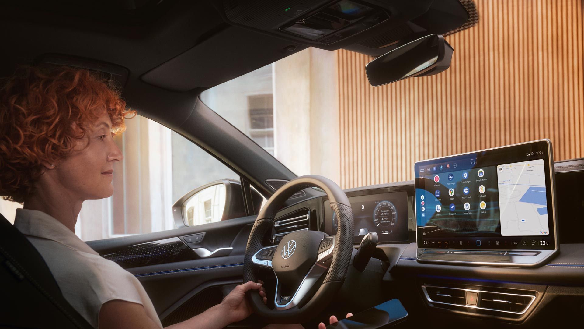 Žena sedí za volantom vozidla VW Tiguan. V ruke drží smartfón a pozerá sa na displej systému infotainment. Druhou rukou drží volant. Cez čelné sklo vidieť moderný dom.