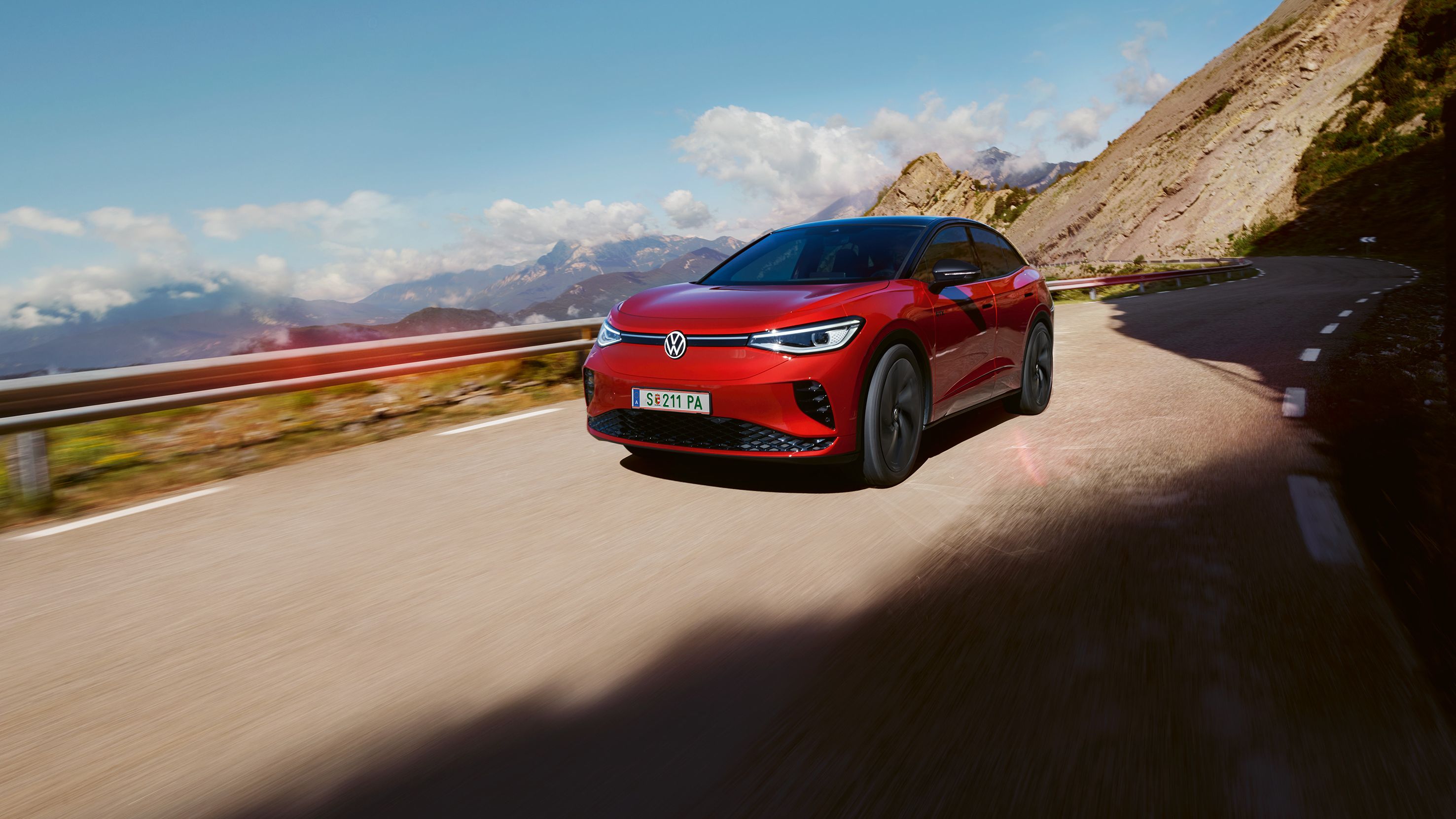 Červené Volkswagen ID.5 GTX  jazdí po ceste v horách. V pozadí vidieť ďalšie kopce zahalené do oblakov.
