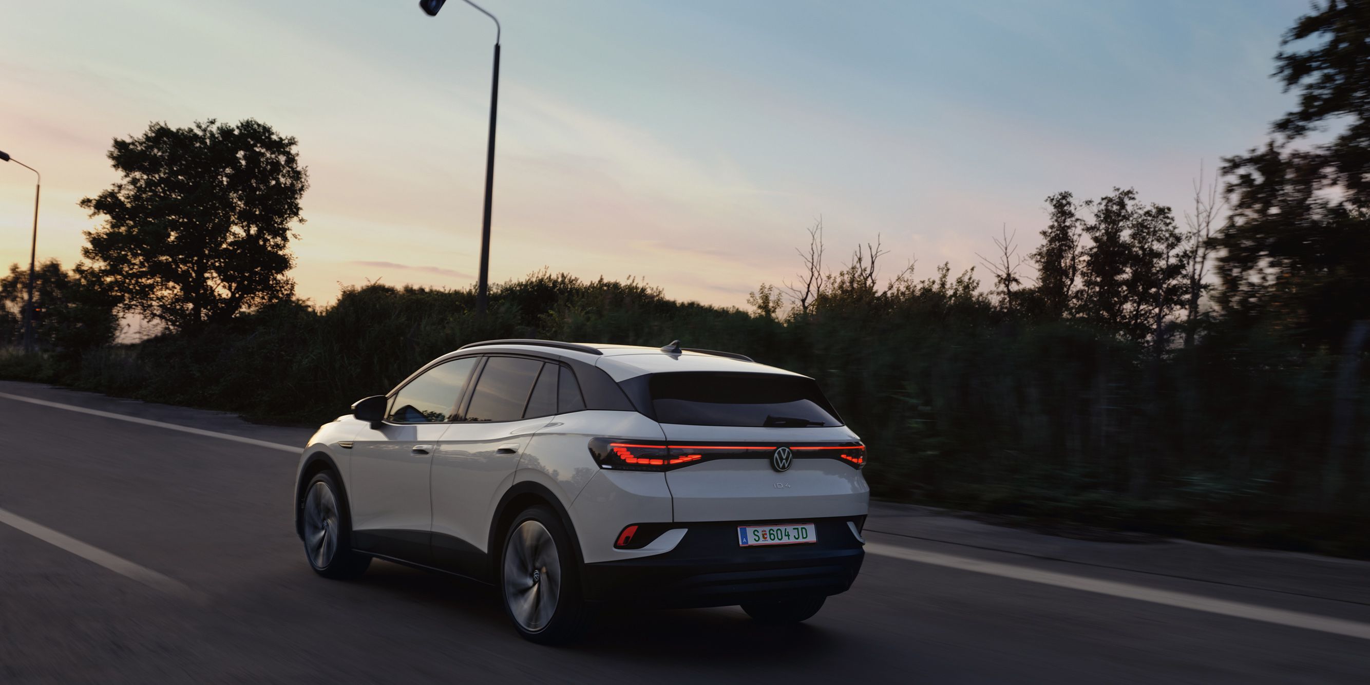 Biely elektrický Volkswagen ID.4 jazdí po ceste za súmraku. Okraj cesty lemujú stromy a kríky. Cesta je rovná a osvetlená pouličnými lampami.