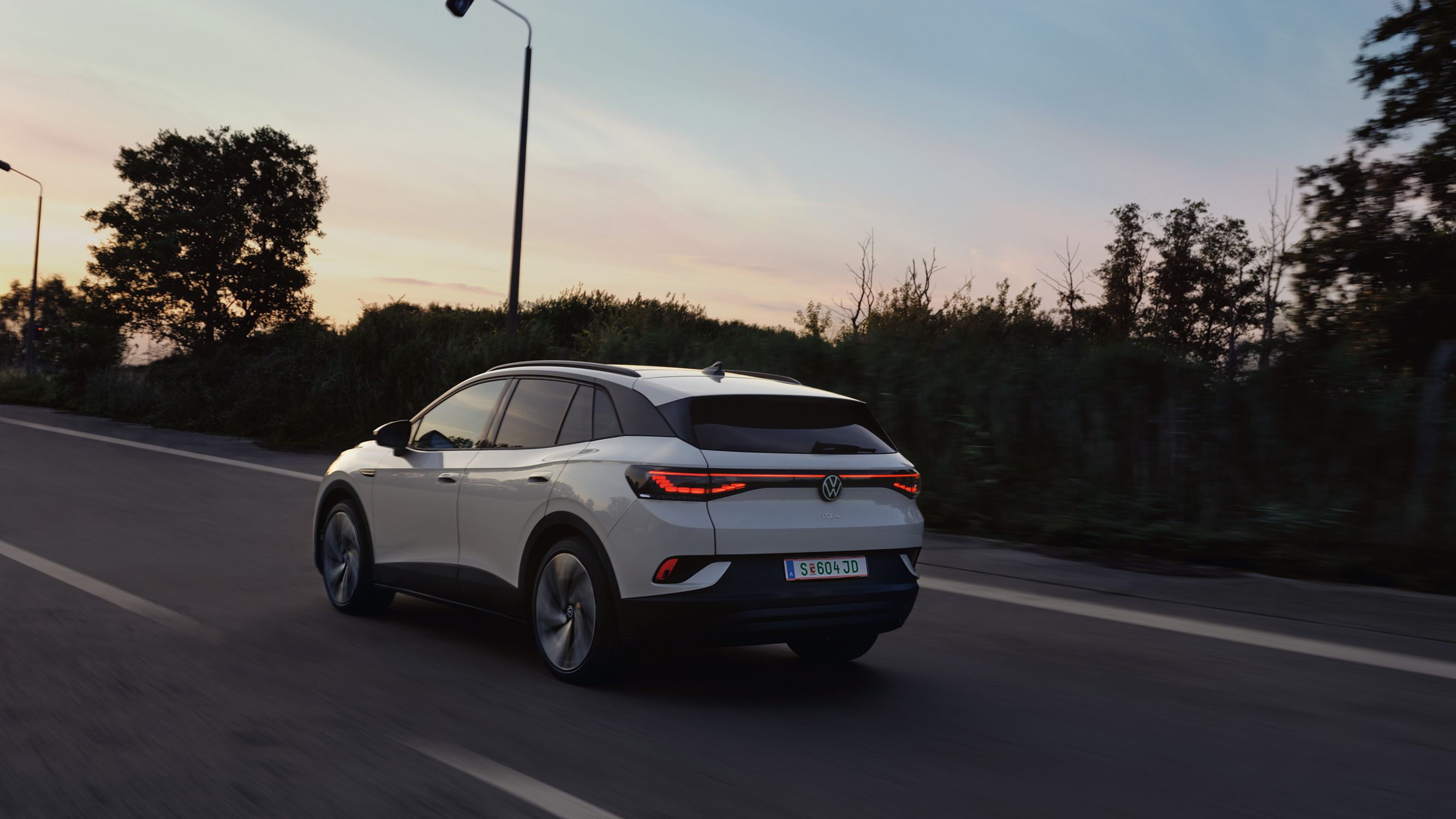 Biely elektrický Volkswagen ID.4 jazdí po ceste za súmraku. Okraj cesty lemujú stromy a kríky. Cesta je rovná a osvetlená pouličnými lampami.