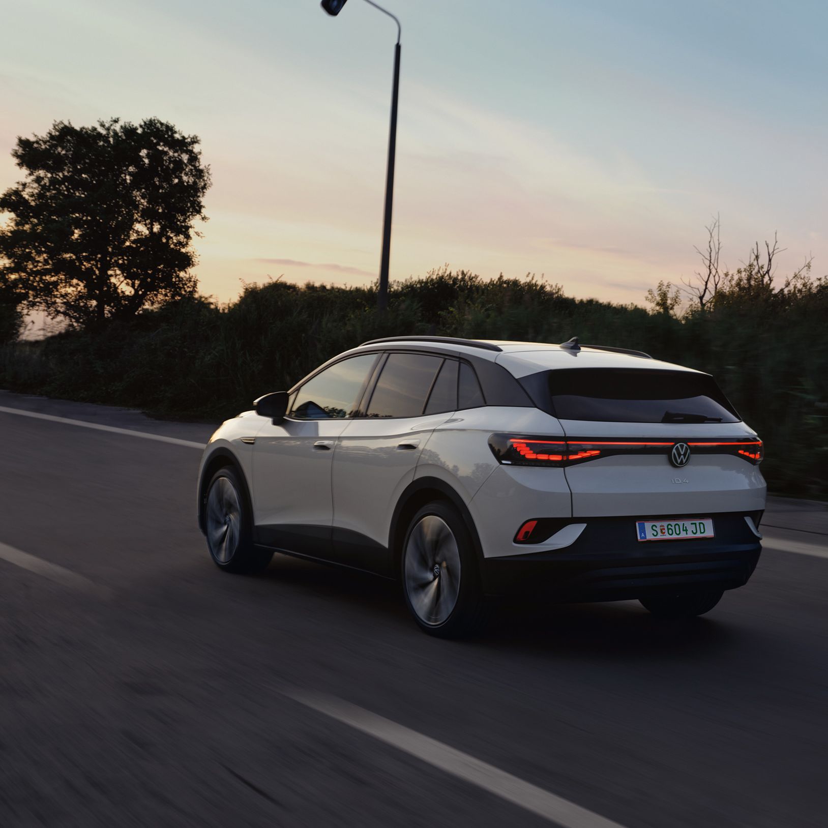 Biely elektrický Volkswagen ID.4 jazdí po ceste za súmraku. Okraj cesty lemujú stromy a kríky. Cesta je rovná a osvetlená pouličnými lampami.