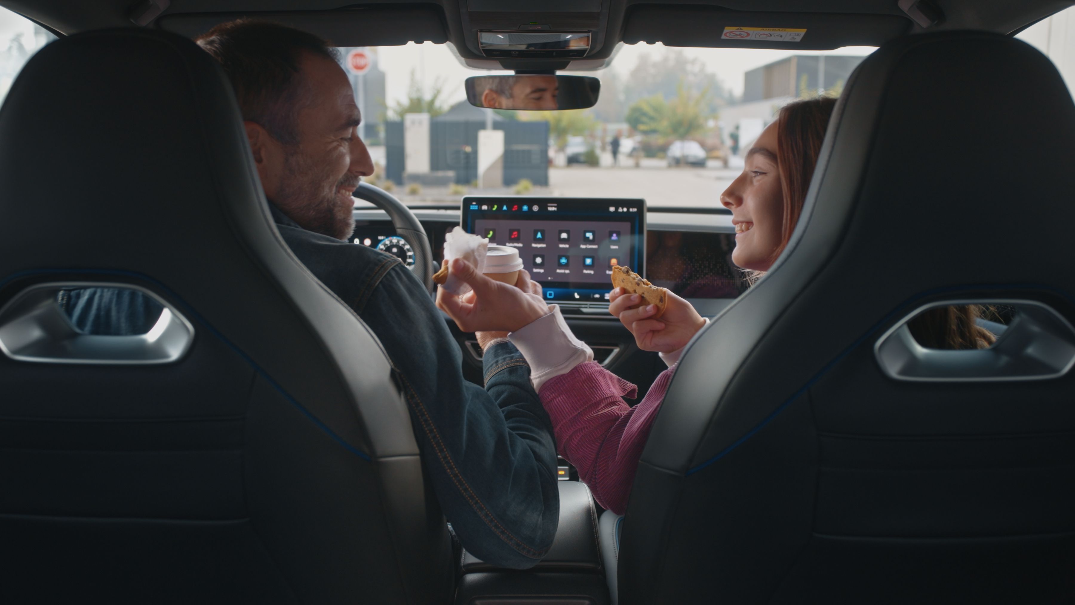 Dve osoby sedia v aute a zdieľajú jedlo. Medzi nimi je viditeľný dotykový displej s rôznymi aplikáciami. Scéna sa odohráva za jasného denného svetla.