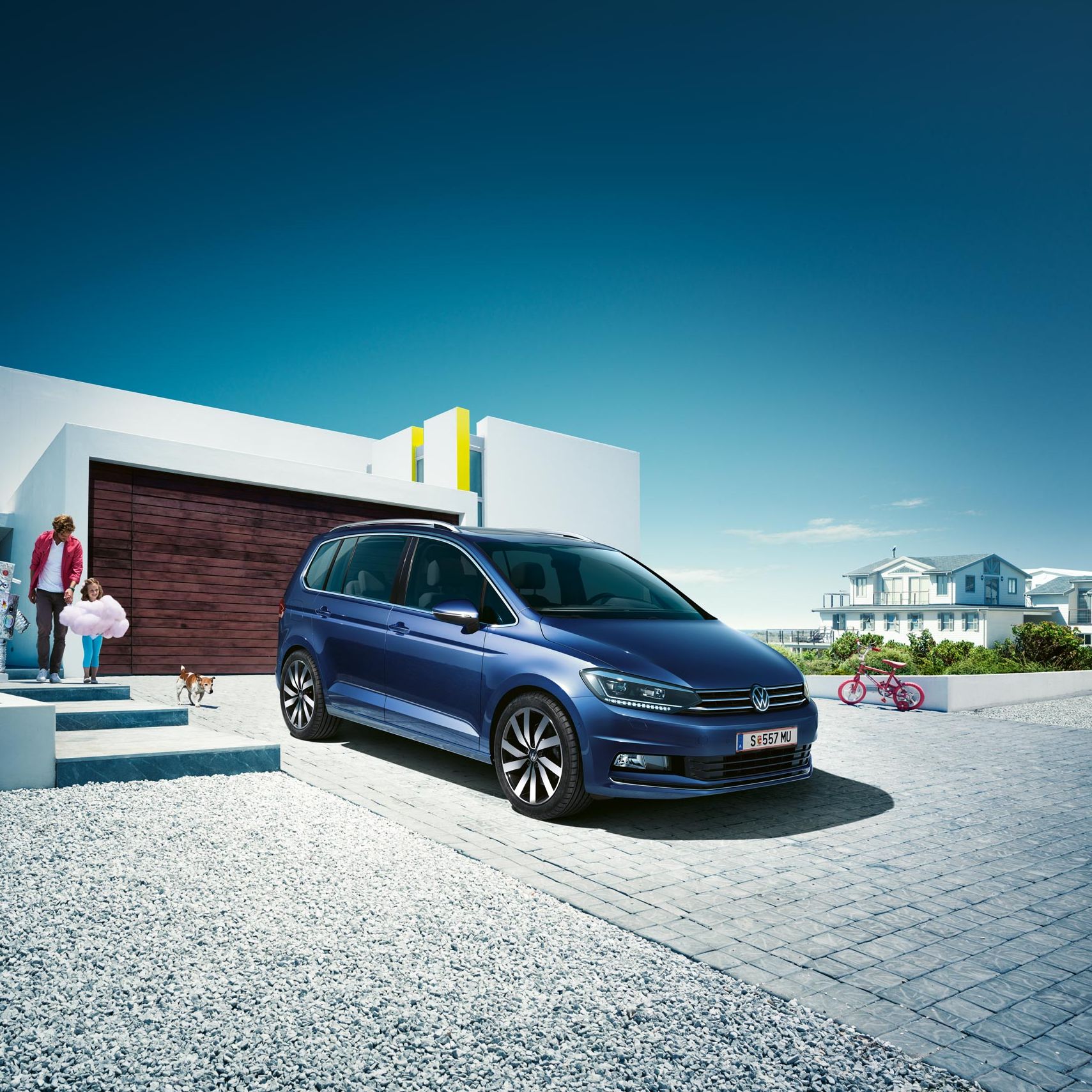 Modrý VW Touran zaparkovaný pred rodinným domom. Z domu vychádza otec s dvomi deťmi, ktoré sú oblečené do kostýmov. Pri nízkom bielom  múriku stojí ružový detský bicykel. V pozadí vidieť ďalšiu veľkú vilu. Obrázku dominuje modrá čistá obloha.