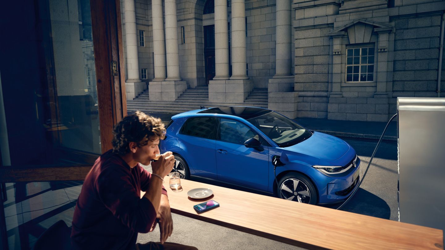 Muž sedí v kaviarni a cez otvorené okno je vidieť modrý VW ID. Polo Style, ktorý sa nabíja na nabíjacej stanici.