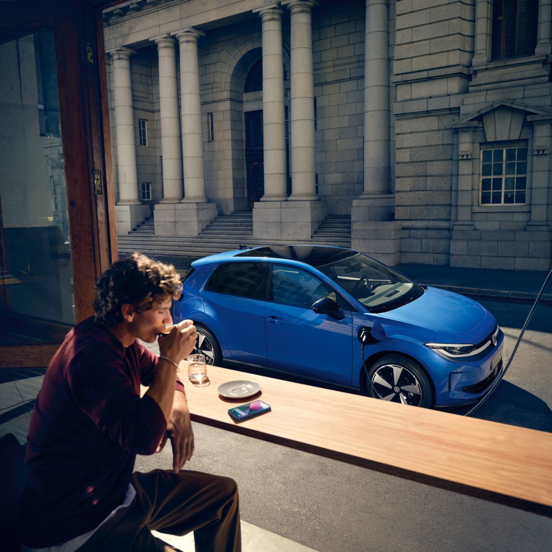 Muž sedí v kaviarni a cez otvorené okno je vidieť modrý VW ID. Polo Style, ktorý sa nabíja na nabíjacej stanici.