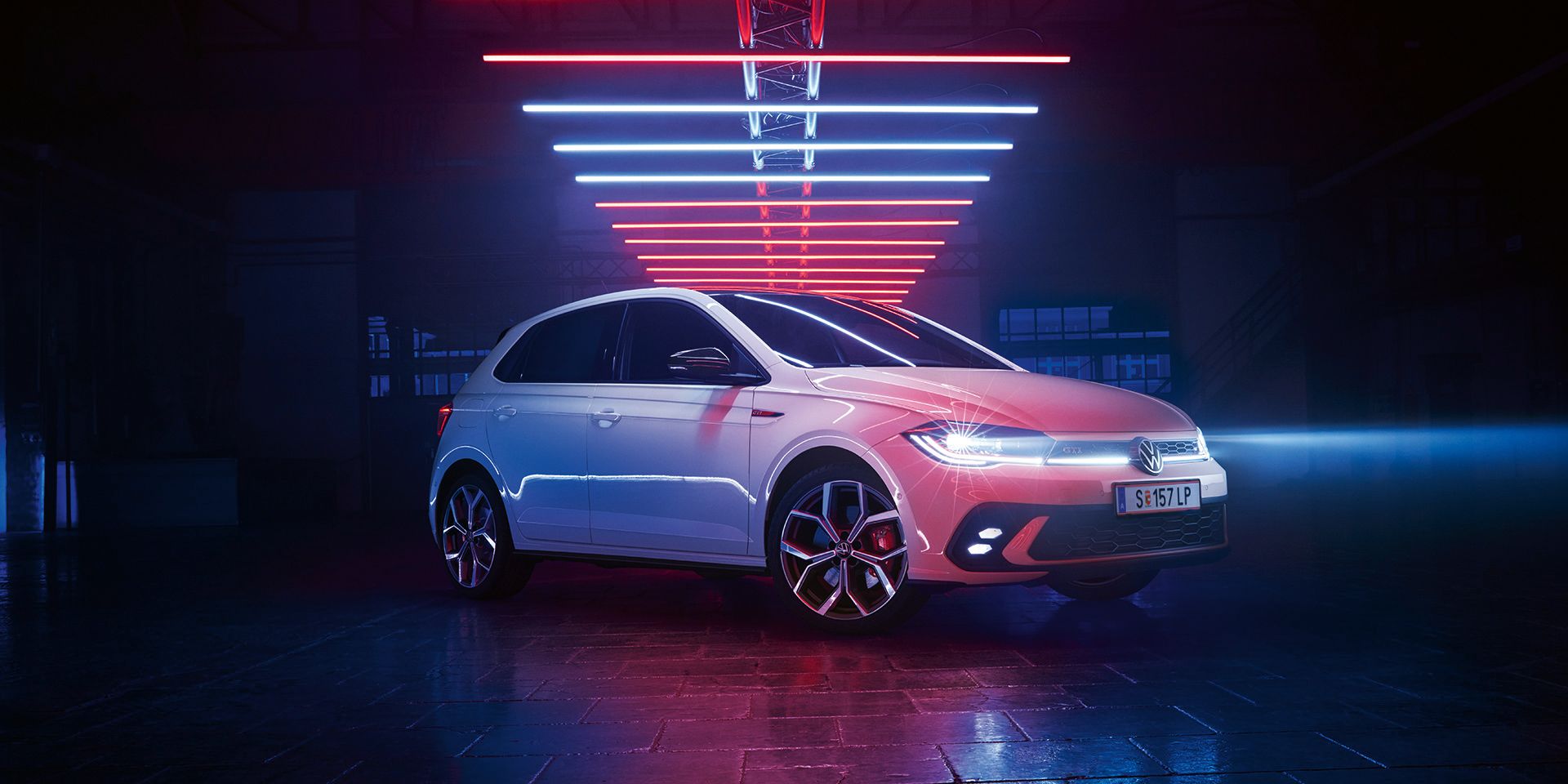 Aufnahme eines weißen VW Polo GTI, der mit angeschalteten LED-Scheinwerfern in einer dunklen Halle unter Leuchtelementen steht.