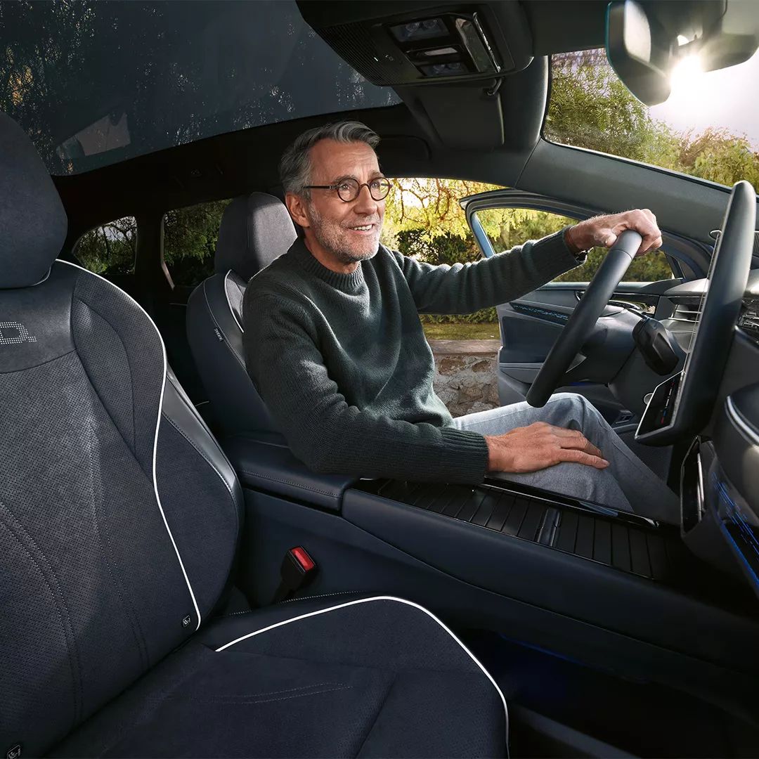 Muž sedí v modernom aute, drží volant a má otvorené dvere. Interiér auta je elegantný s pohodlnými sedadlami. Von vidieť prírodu.