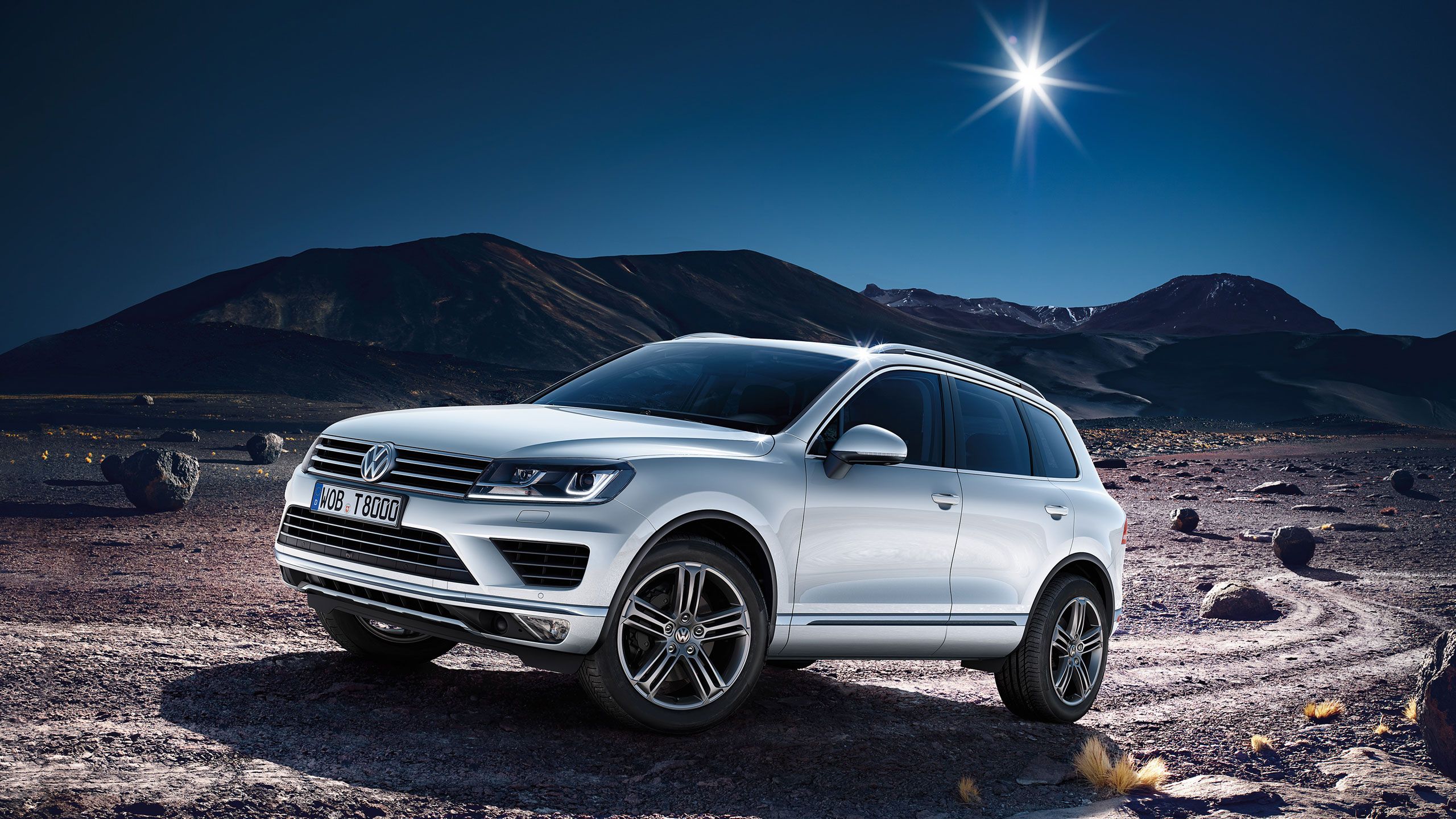 Na obrázku je biely SUV automobil značky Volkswagen zaparkovaný na nehostinnej hornatej krajine počas večera, keď je na oblohe jasná hviezda. Automobil má moderný a športový vzhľad. V pozadí sú tmavé hory a skalnaté útvary.