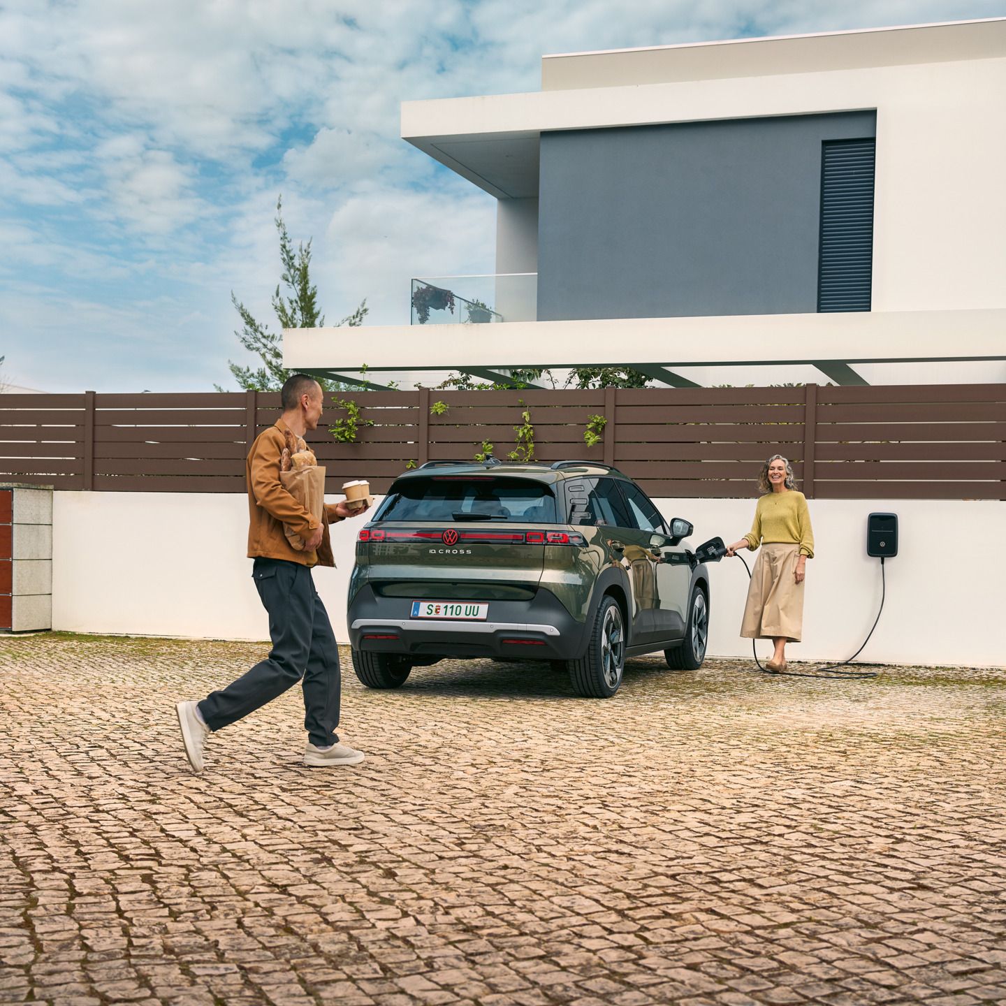 Žena zapája nabíjací kábel zo zásuvky do zeleného VW ID. Cross, muž, k nej pristúpi s kávou so sebou v ruke a pečivom.