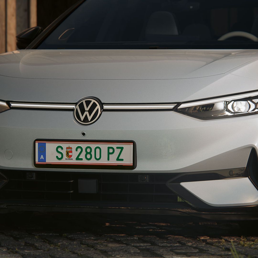 Elegantné biele auto značky Volkswagen s rakúskou poznávacou značkou stojí na dlažobných kockách pred drevenou stavbou. Predné svetlomety vozidla sú zapnuté.