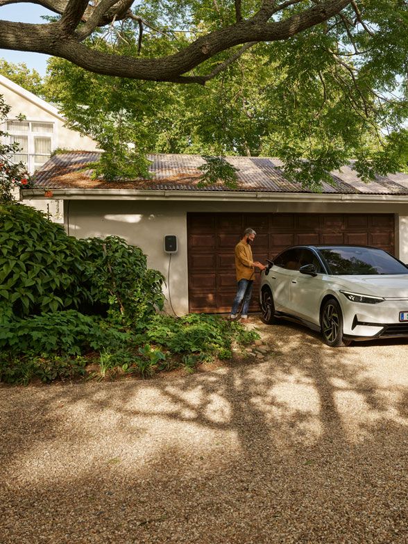 Muž stojí vedľa bieleho elektrického auta značky Volkswagen zaparkovaného pred garážou v záhrade plnej zelene. Okolo auta sú veľké stromy a rôzne kvitnúce rastliny. Garáž má hnedé dvere.
