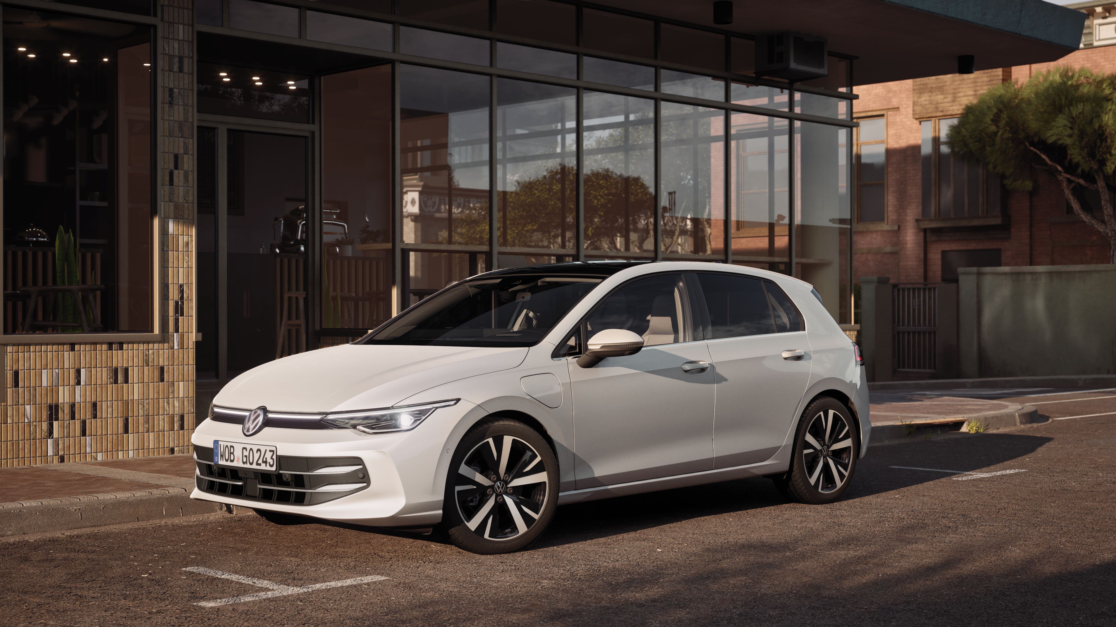 Biely hatchback Volkswagen zaparkovaný pred modernou budovou s čiastočne sklenenou fasádou. Auto stojí na slnečnej ulici s dláždeným chodníkom. Vozidlo má elegantný dizajn a športové disky z ľahkej zliatiny.