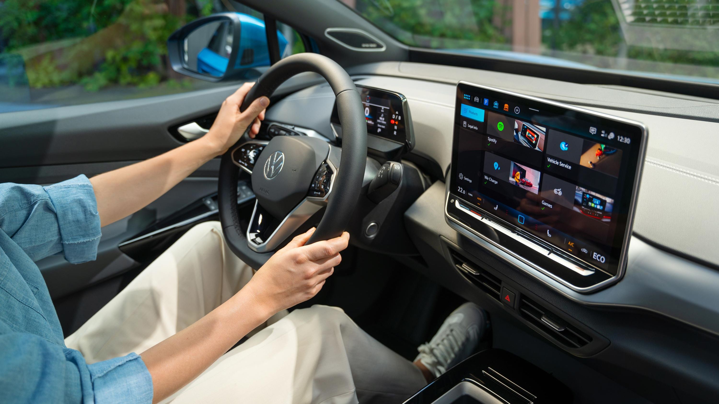 Pohľad do interiéru VW ID.4. Bočný pohľad na multifunkčný volant a systém infotainment. Osoba sediaca na mieste vodiča riadi vozidlo. 