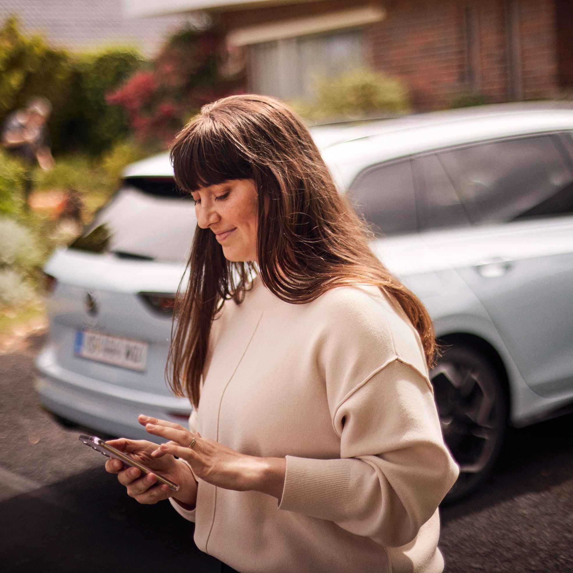 Žena stojí za vozidlom Volkswagen a pozerá na svoj smartfón, ktorým niečo ovláda. Vozidlo je rozostrené a stojí pred domom. Za vozidlom je krásna záhrada.