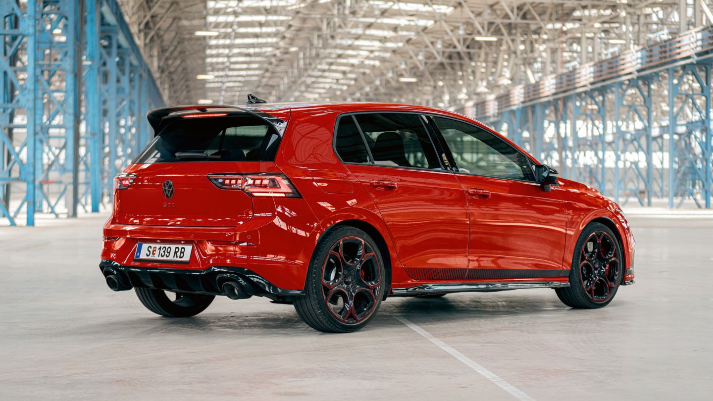 Červený VW Golf GTI je zaparkovaný vo veľkej, svetlom zaliatej priemyselnej hale. Auto je viditeľné zozadu a mierne zboku. Čierne kolesá a zadný spojler sú jasne viditeľné.