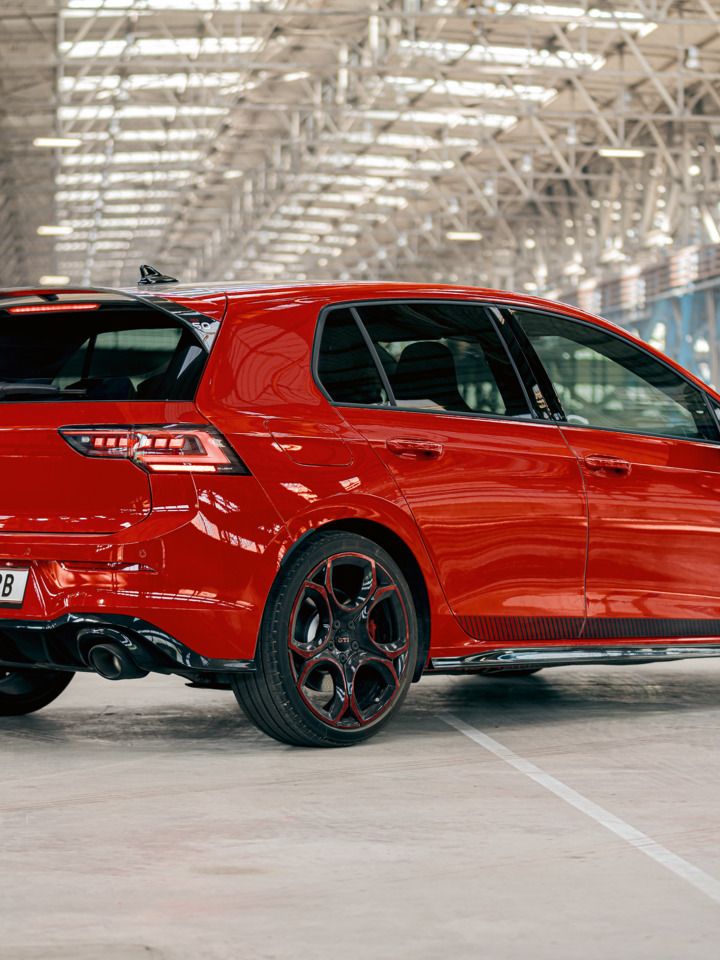 Červený VW Golf GTI je zaparkovaný vo veľkej, svetlom zaliatej priemyselnej hale. Auto je viditeľné zozadu a mierne zboku. Čierne kolesá a zadný spojler sú jasne viditeľné.