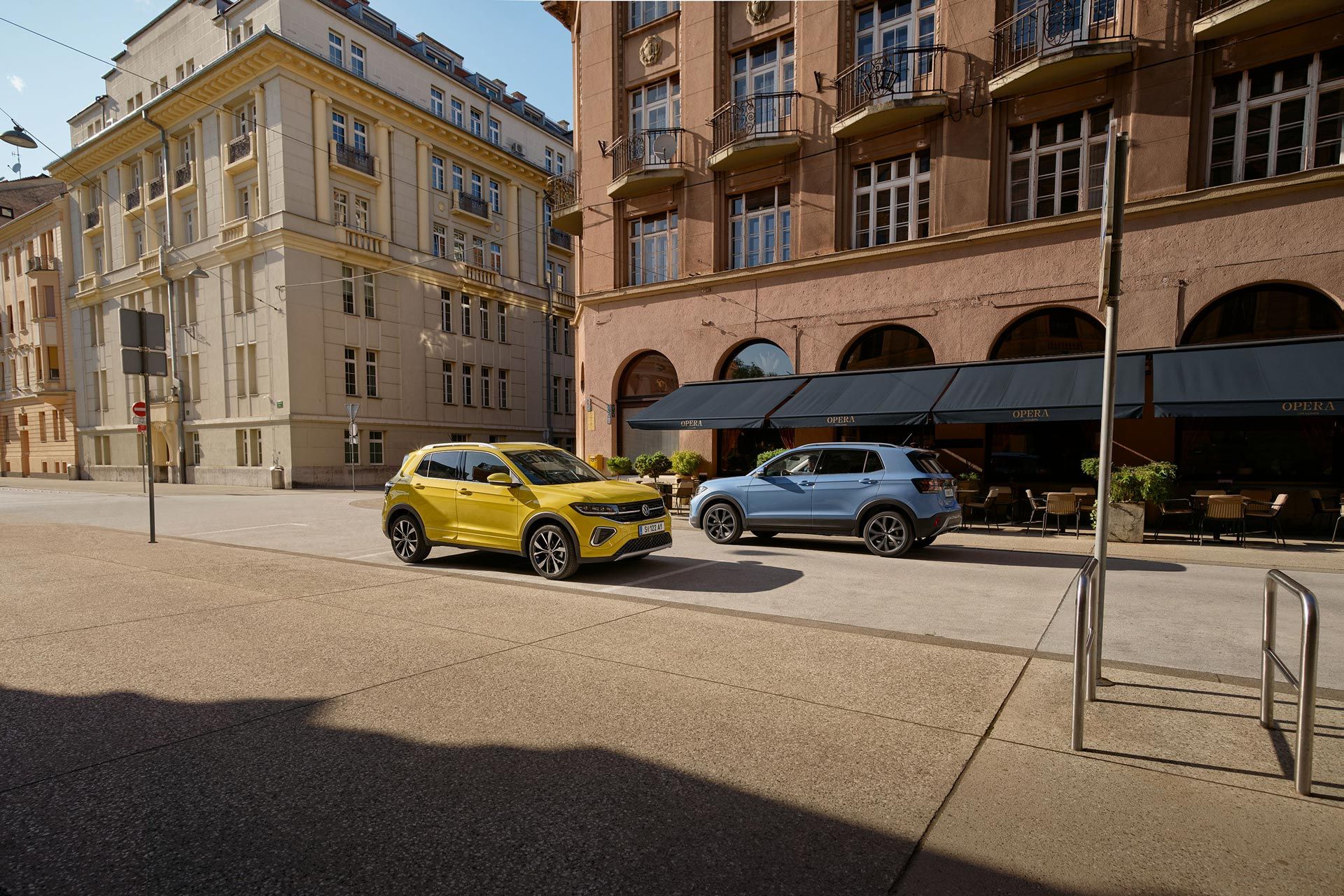 Po ceste v centre mesta prechádzajú okolo seba žltý a modrý VW T-Cross. Za nimi vidieť historické budovy.