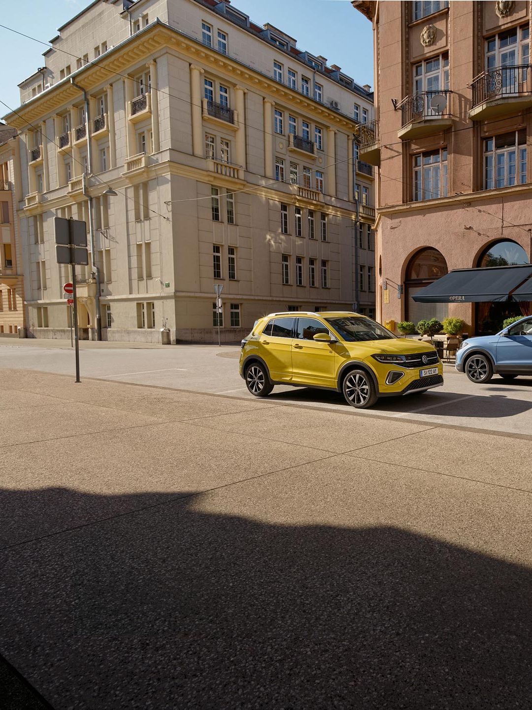 Po ceste v centre mesta prechádzajú okolo seba žltý a modrý VW T-Cross. Za nimi vidieť historické budovy.