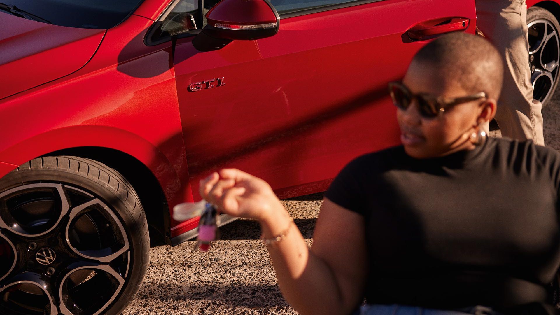 Detailný záber na predné partie červeného VW Golf GTI zboku so ženou v popredí, ktorá sedí na stoličke pred automobilom. Muž za ňou stojí v otvorených dverách vodiča. Vozidlo stojí na štrkovej ceste.