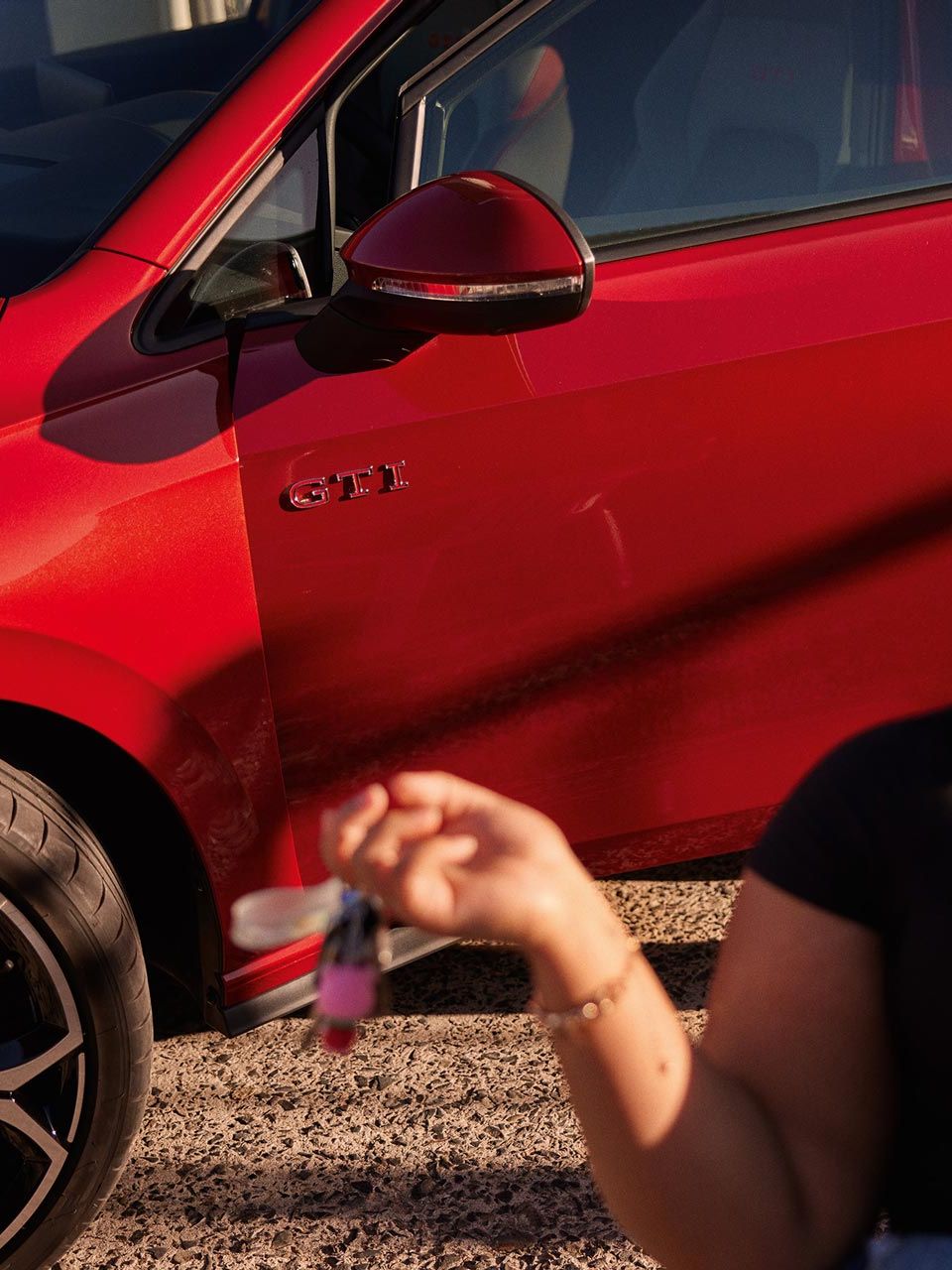 Detailný záber na predné partie červeného VW Golf GTI zboku so ženou v popredí, ktorá sedí na stoličke pred automobilom. Muž za ňou stojí v otvorených dverách vodiča. Vozidlo stojí na štrkovej ceste.
