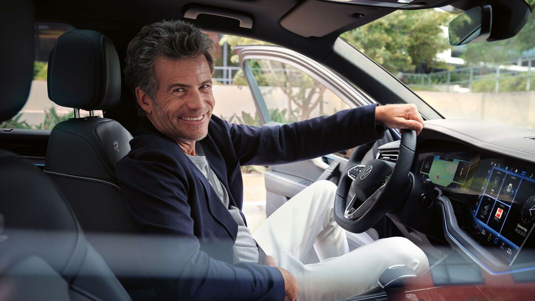 Usmiaty muž sedí  na mieste vodiča v modeli Touareg s rukou opretou o volant. Moderný interiér vozidla disponuje dotykovou obrazovkou a digitálnymi prvkami.