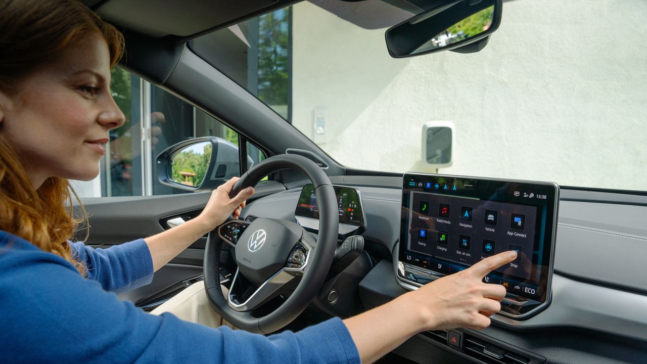 Žena sedí v aute a dotýka sa dotykového displeja na palubnej doske. Displej zobrazuje rôzne aplikácie a informácie. Volant auta má logo Volkswagen a rôzne tlačidlá na ovládanie funkcií vozidla.