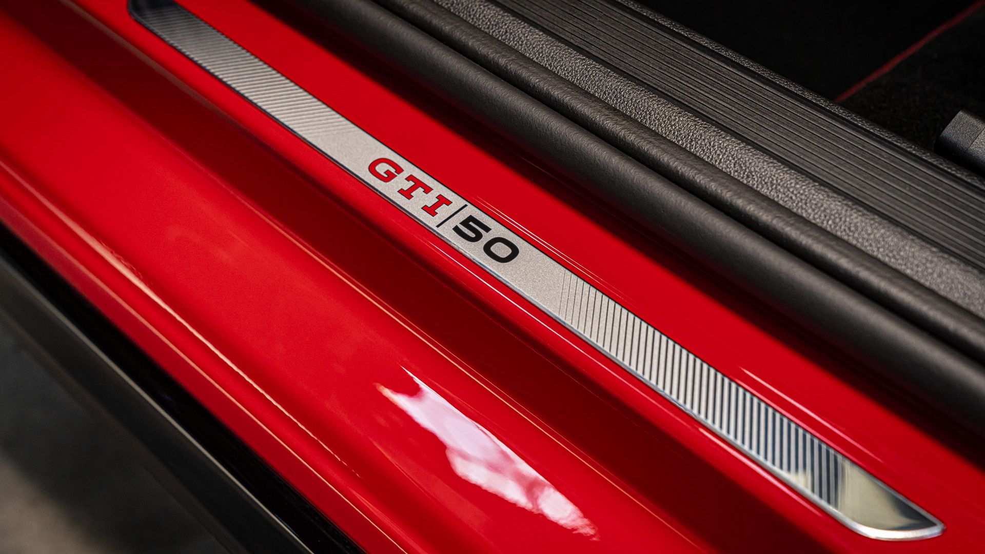 Detailný záber na prah dverí červeného modelu Golf GTI so striebornou plaketou. Na plakete je nápis „GTI 50“. Obrázok zobrazuje detailné spracovanie a športový dizajn vozidla.