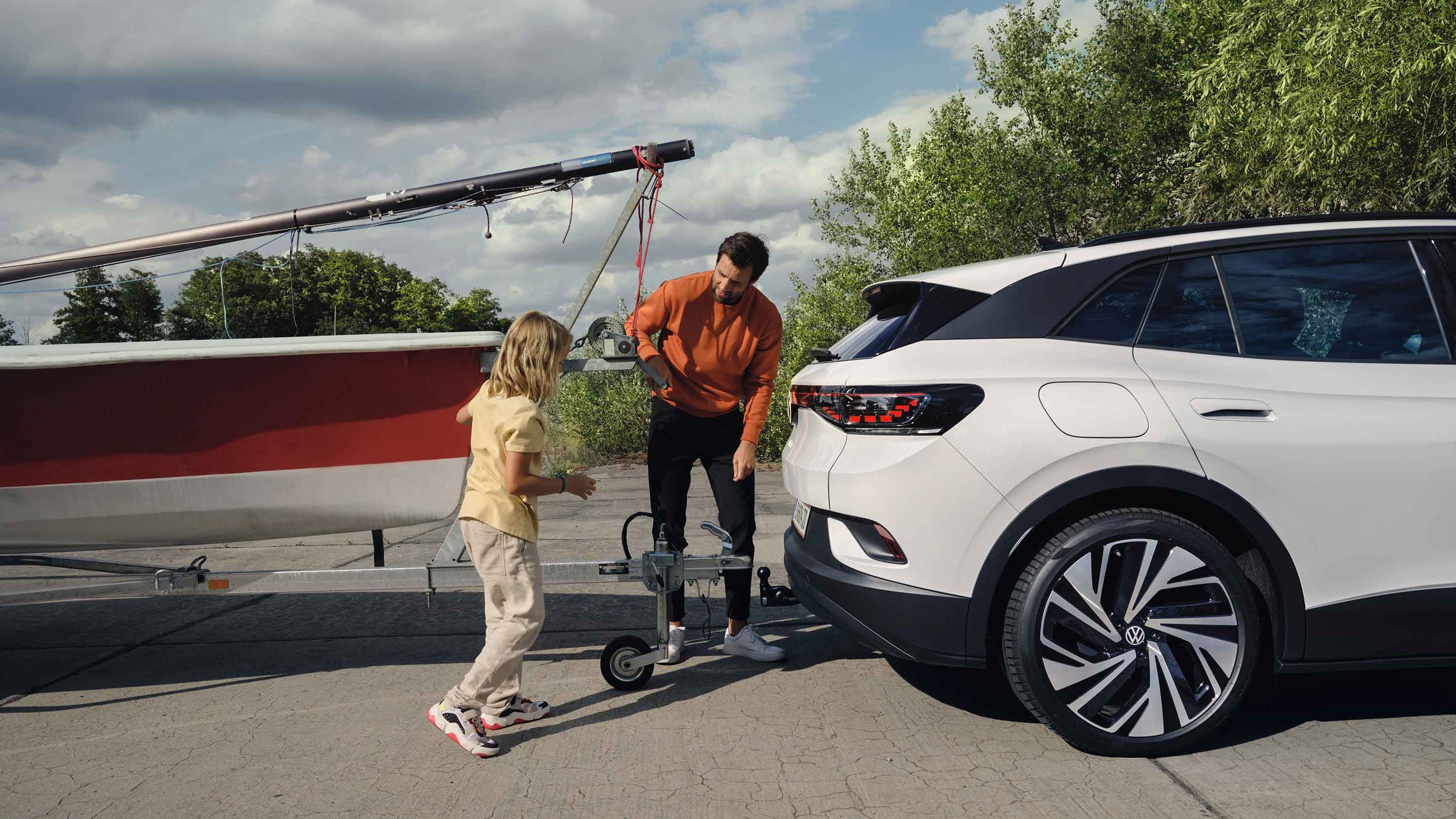 Muž a dievča zapájajú prívesný vozík s loďou k ťažnému zariadeniu elektrického SUV. Okolo nich je zeleň s niekoľkými stromami a čiastočne zamračenou oblohou.