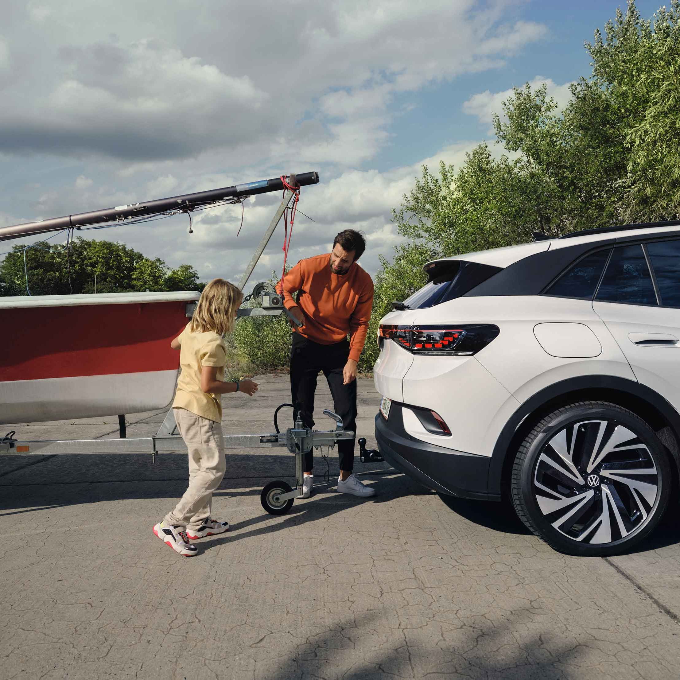 Muž a dievča zapájajú prívesný vozík s loďou k ťažnému zariadeniu elektrického SUV. Okolo nich je zeleň s niekoľkými stromami a čiastočne zamračenou oblohou.