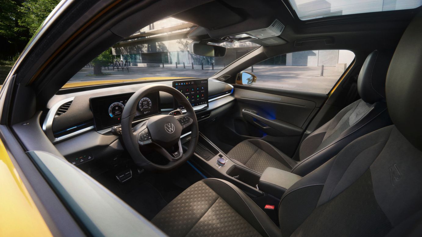 Obrázok ukazuje pohľad do interiéru žltého VW T-Roc. Vidieť predné sedadlá, volant, infotainment systém, ale aj malú časť žltého exteriéru. V pozadí je vidieť mestskú budovu a vedľa nej husté listnaté stromy.