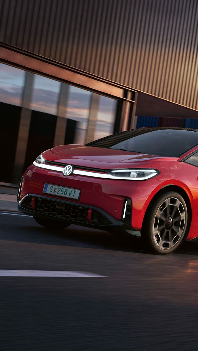 [AI-modified] – Červený VW ID.3 GTI prechádza okolo prístavnej budovy so zapnutými svetlometmi a rozsvieteným logom.