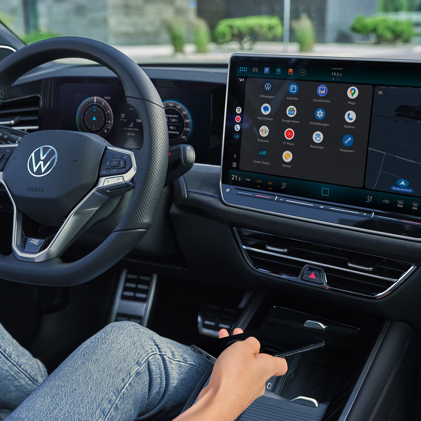 Detailný pohľad na displej systému infotainment v VW Passast. Na mieste vodiča sedí žena v ruke drží smartfón, ktorý ukladá do priehradky na nabíjanie. Cez čelné sklo vidieť mestské prostredie.