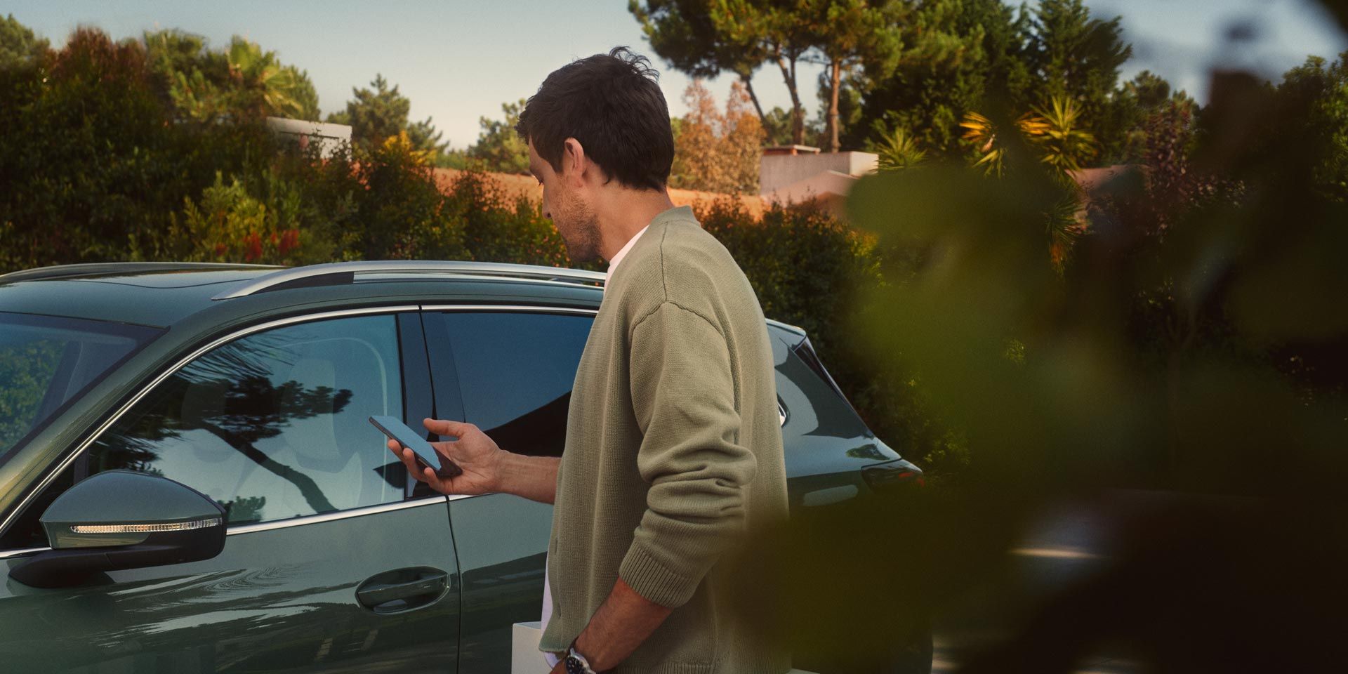 Mladý muž stojí na strane vodiča tmavozeleného VW Tiguan a pomocou smartfónu ovláda systém Park Assist Plus.