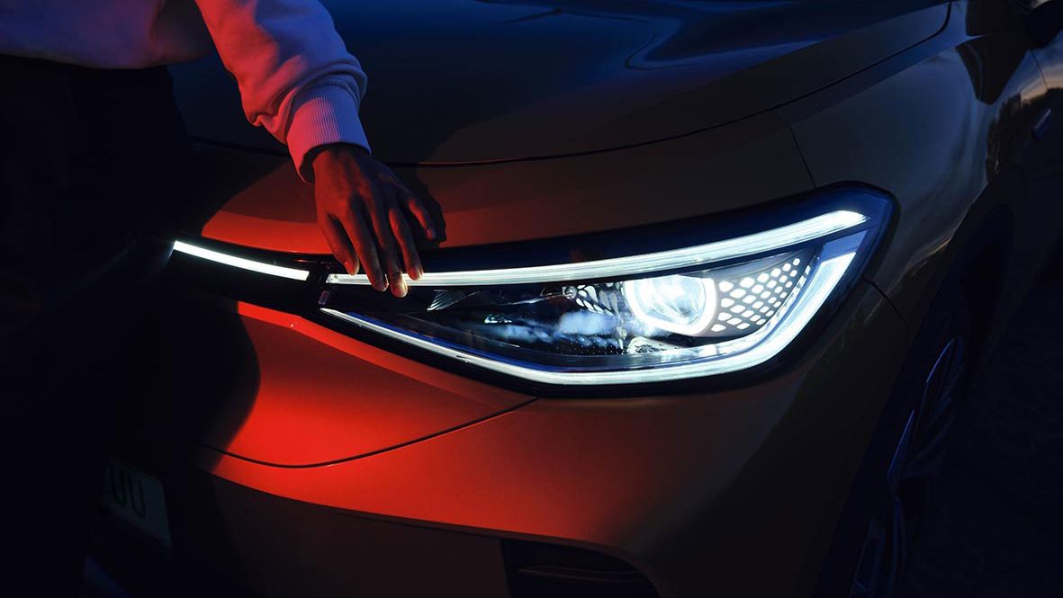 Osoba sa dotýka predného zasvieteného svetlometu v tme. Svetlo automobilu je jasné a detailne prepracované. Fotografiu zdôrazňuje kontrast medzi svetlom a tmavým prostredím.
