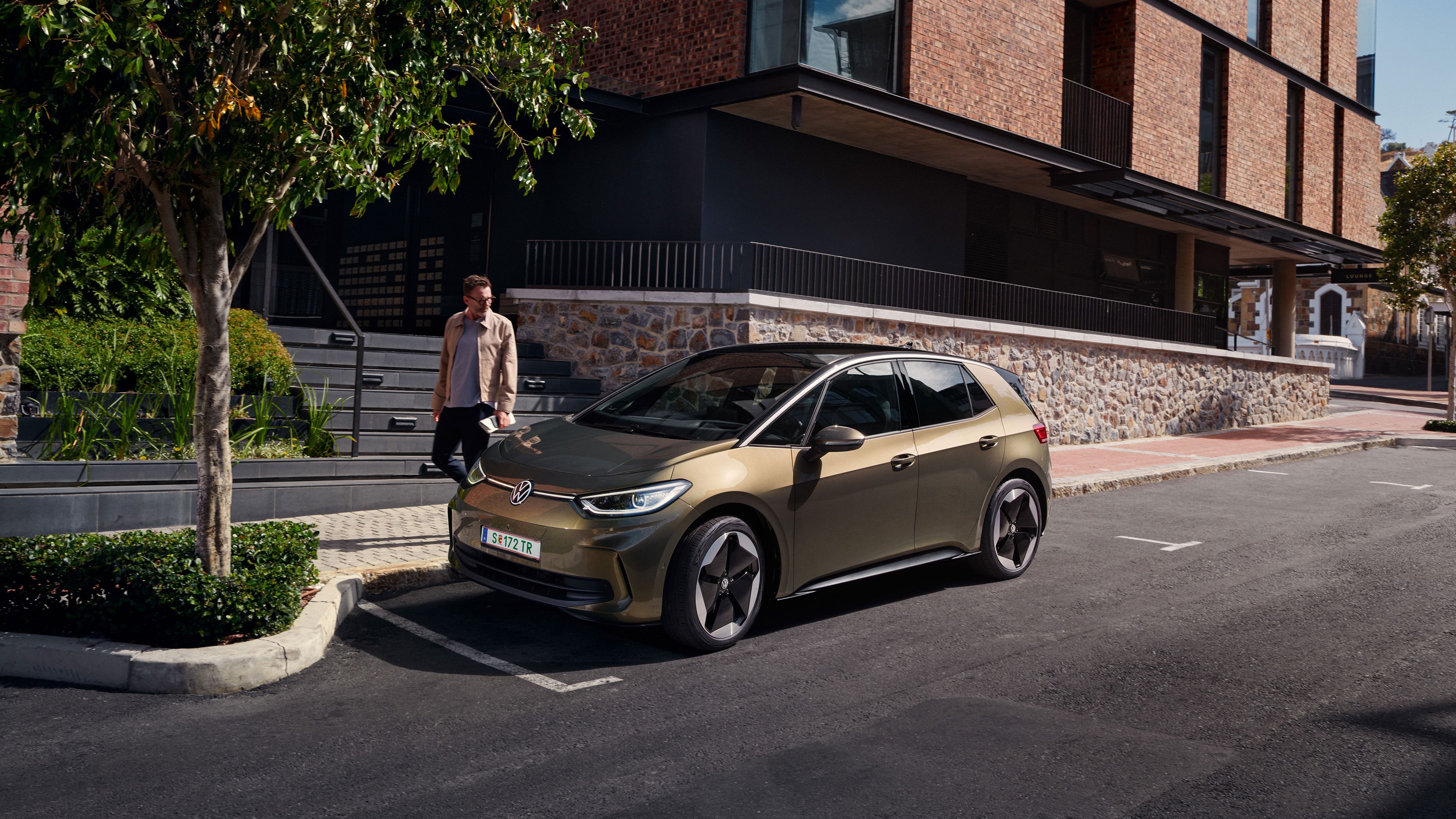 Elektrické vozidlo značky Volkswagen stojí na parkovacom mieste pri chodníku pred moderným bytovým domom. Okolo auta prechádza muž po chodníku. Pri chodníku rastú stromy.
