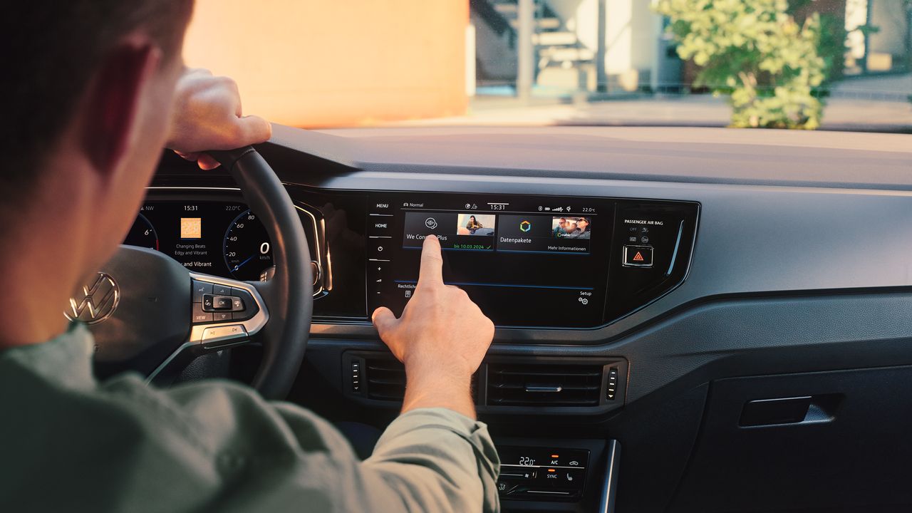 Muž ovláda internetový obchod VW upgrads vo VW Polo prostredníctvom displeja systému infotainment. Čiastočne vidieť volant a digitálny prístrojový panel. Cez čelné sklo vidieť strom a budovu.