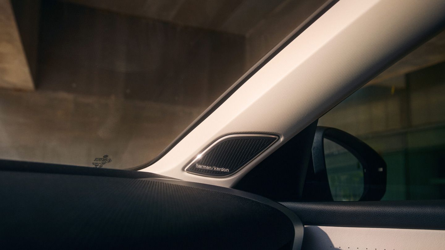 Detailný záber interiéru vozidla T-Roc so zameraním na reproduktor Harman Kardon v stĺpiku A. Vidieť aj časť posuvnej strechy a bočné zrkadlá vozidla.