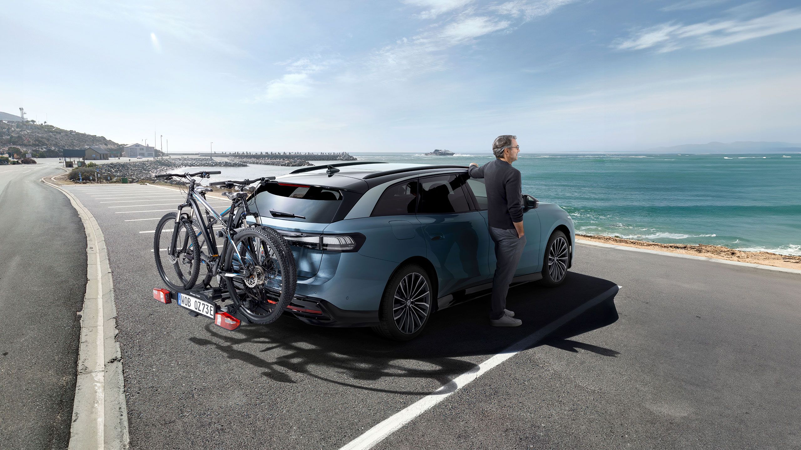 Muž stojí vedľa vozidla ID.7 Tourer s nosičom bicyklov VW a pozerá sa na more.