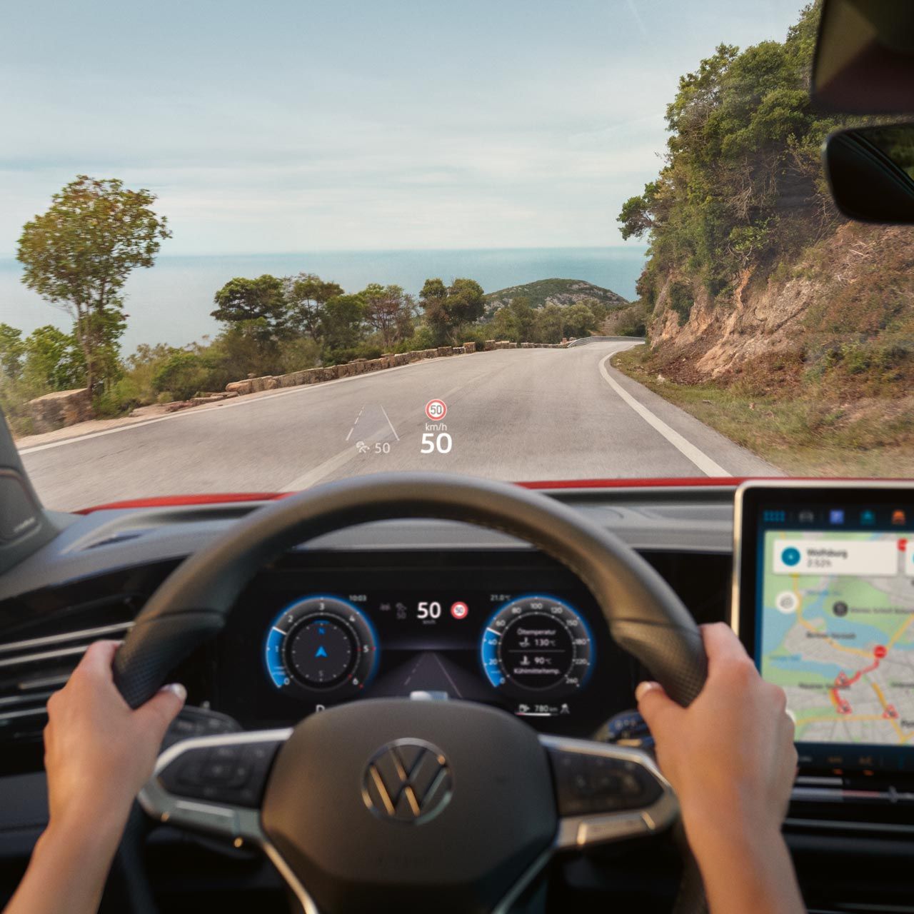 Pohľad do interiéru auta na volant a digitálny prístrojový panel, displej systému infotainment s navigačnou mapou počas jazdy po horskej ceste. Vodič drží volant. Cez čelné sklo vidieť cestu je ohraničená stromami a skalnou stenou.