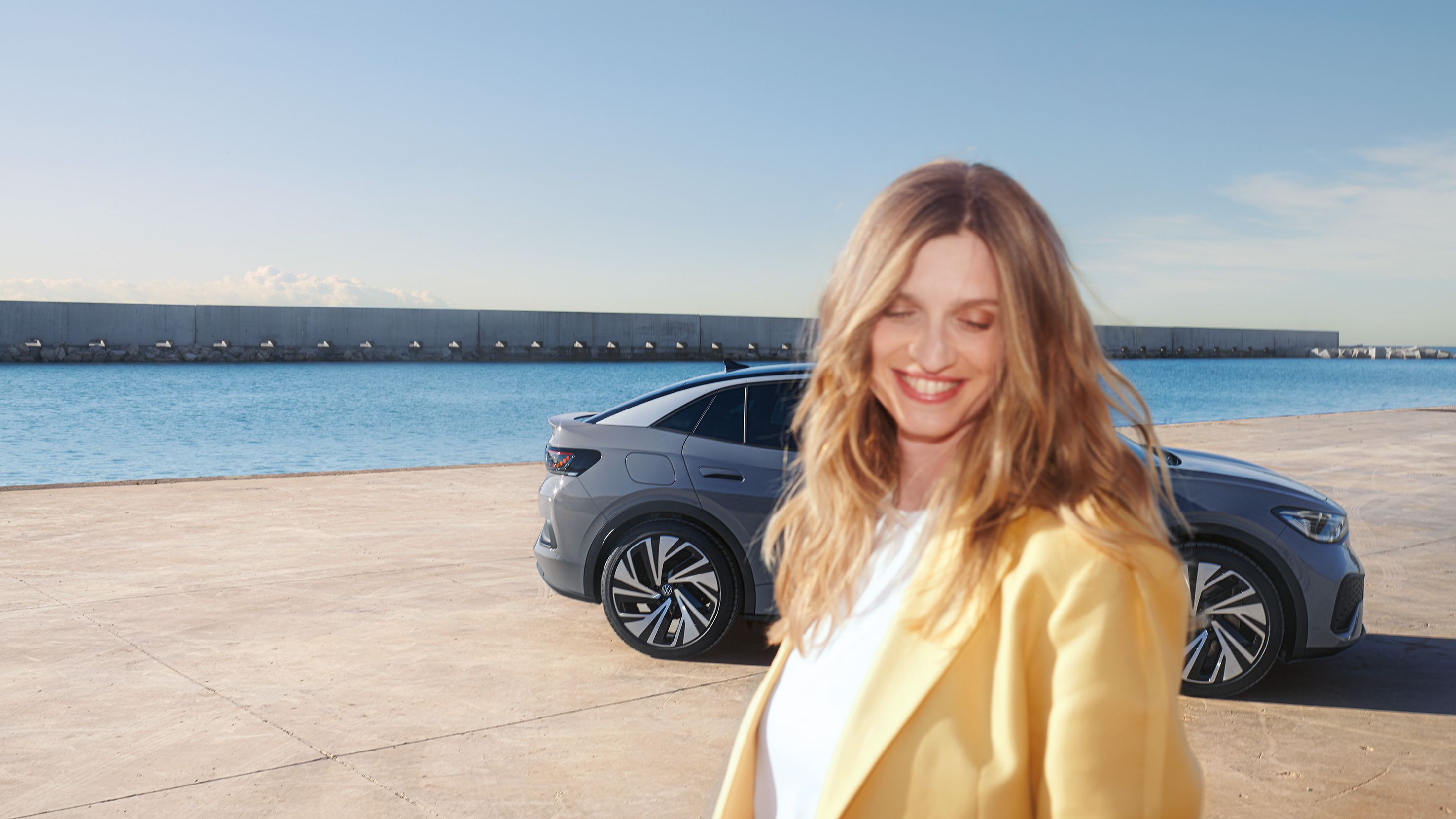 Bočný pohľad na sivý VW ID.5 na nábreží v prístave, v popredí stojí usmievavá, pestro oblečená žena.