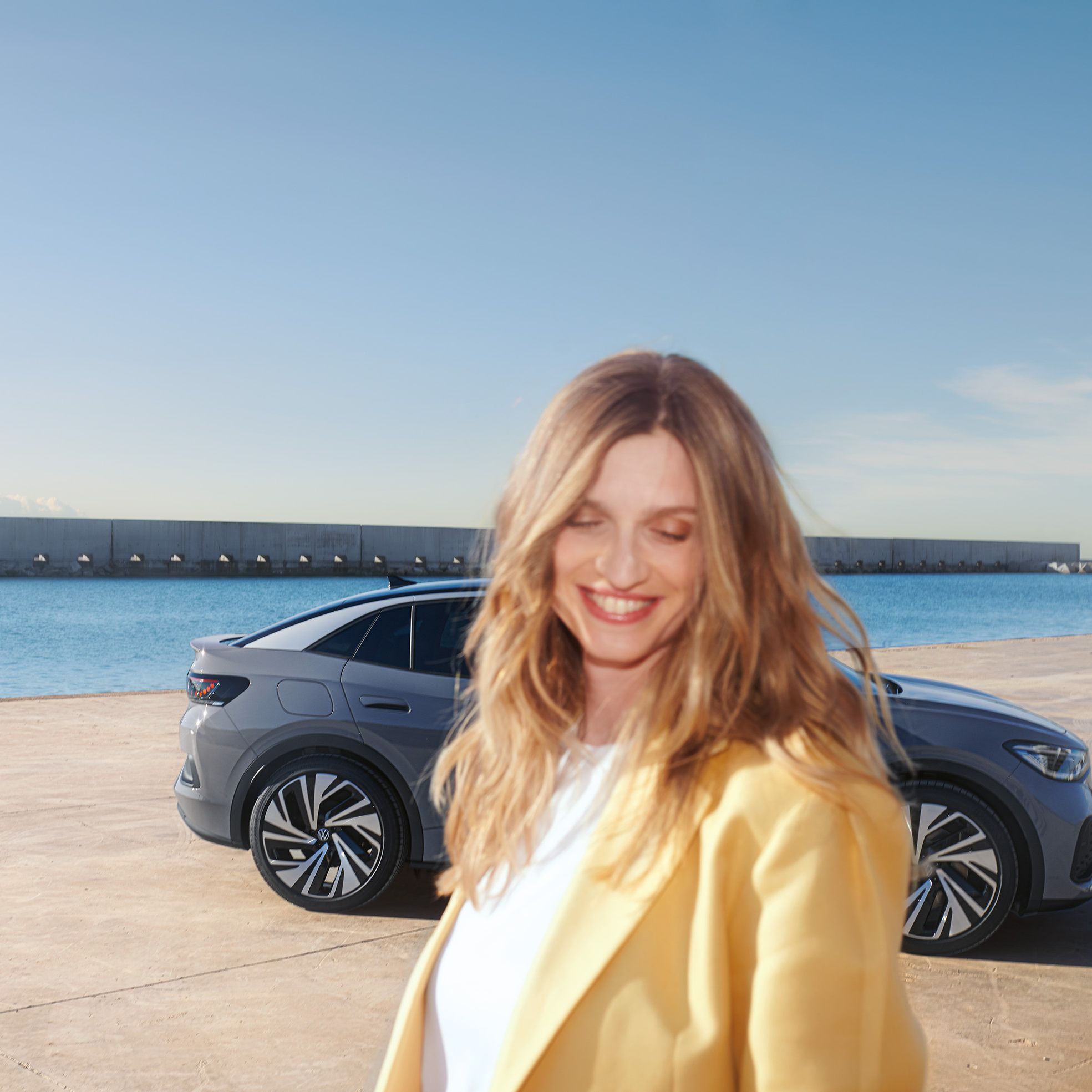 Bočný pohľad na sivý VW ID.5 na nábreží v prístave, v popredí stojí usmievavá, pestro oblečená žena.