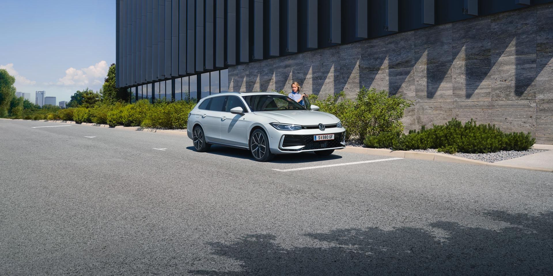 Biele auto zaparkované pred modernou budovou s tmavou fasádou. Žena nastupuje do vozidla na mieste vodiča. Vedľa budovy je zeleň.