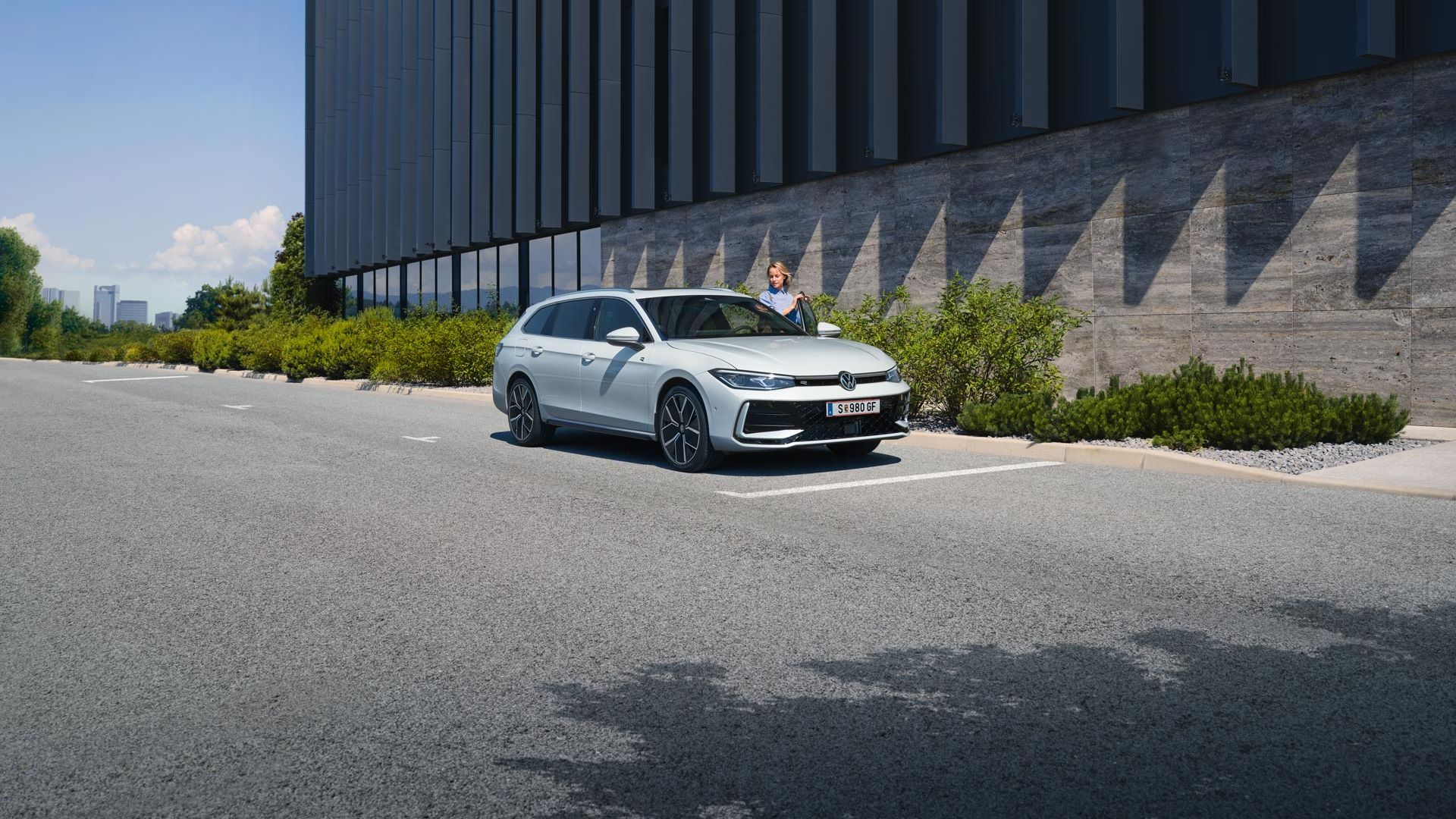 Biele auto zaparkované pred modernou budovou s tmavou fasádou. Žena nastupuje do vozidla na mieste vodiča. Vedľa budovy je zeleň.