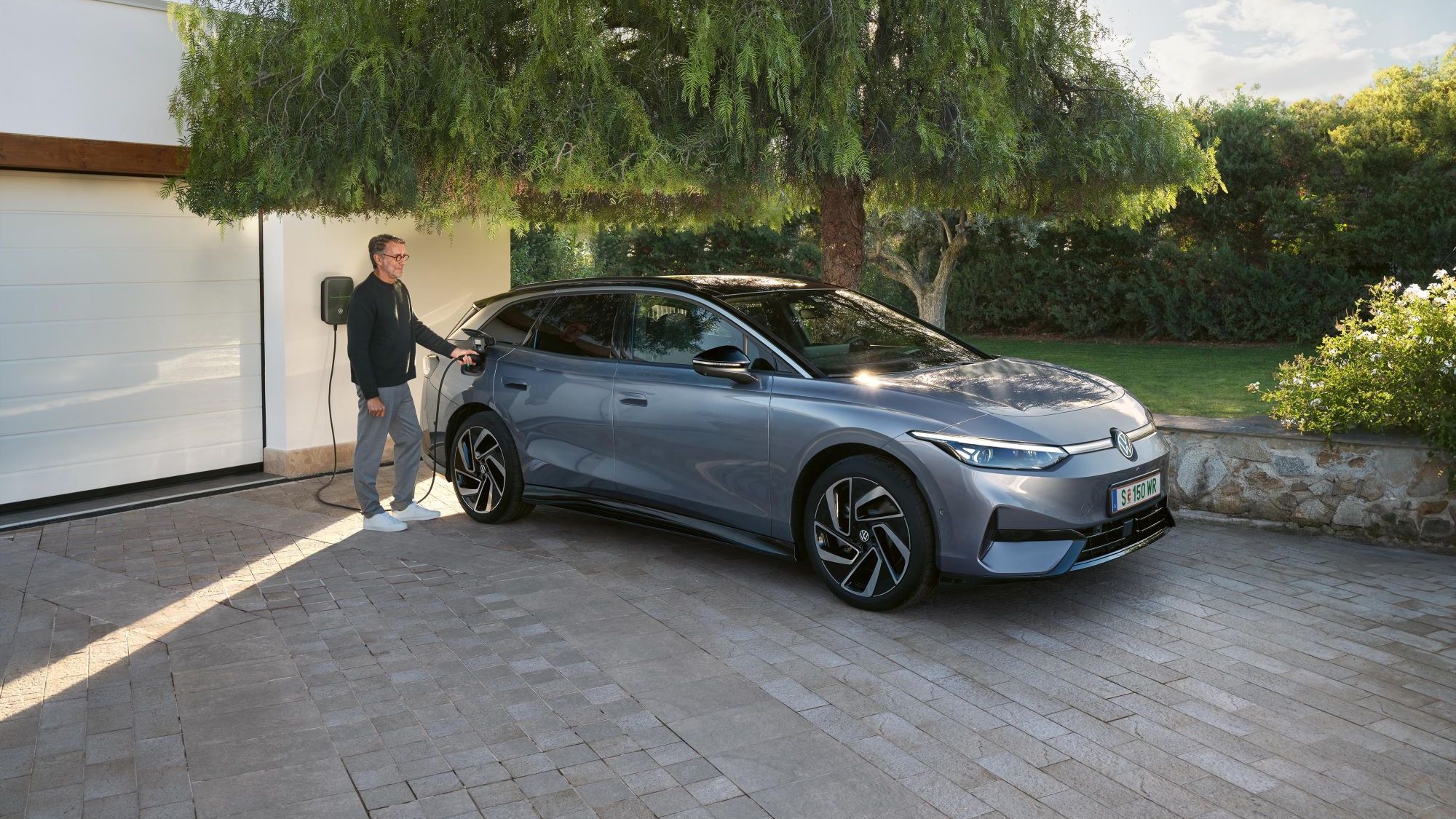 Muž stojí vedľa modrého VW ID.7 Tourer, ktorý sa nabíja pred garážou pri nabíjacej stanici Wallbox.
