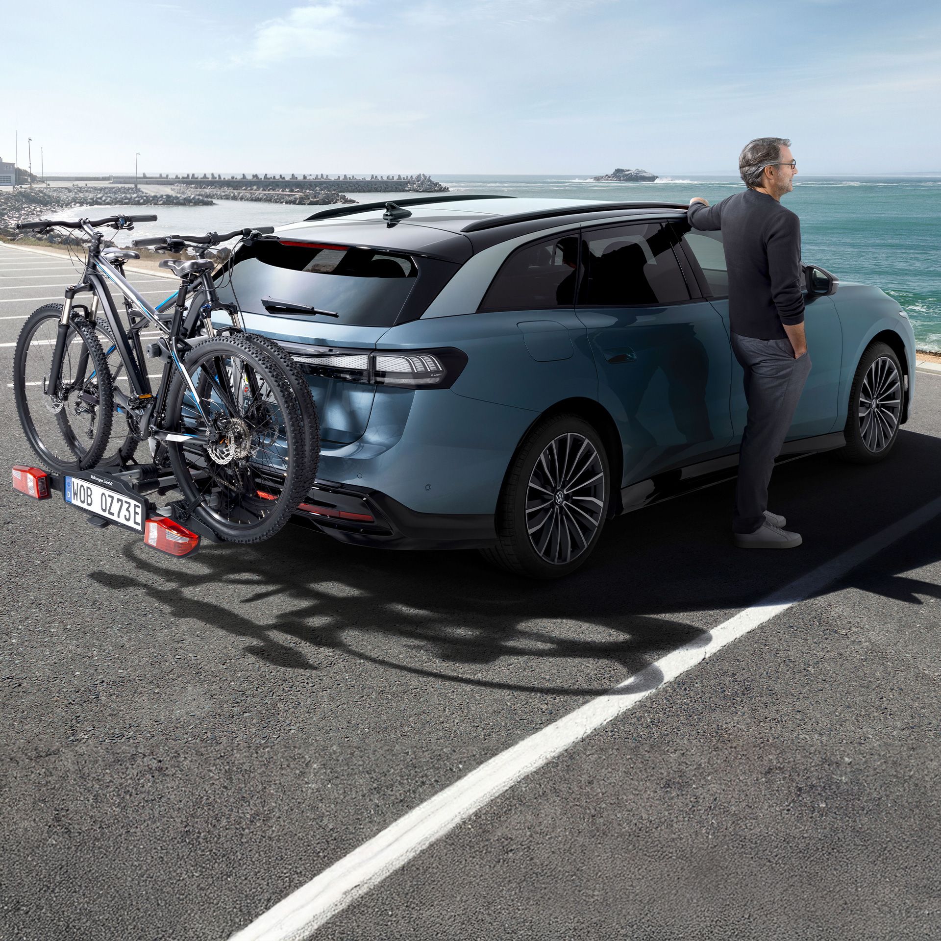 Muž stojí vedľa vozidla ID.7 Tourer s nosičom bicyklov VW a pozerá sa na more.
