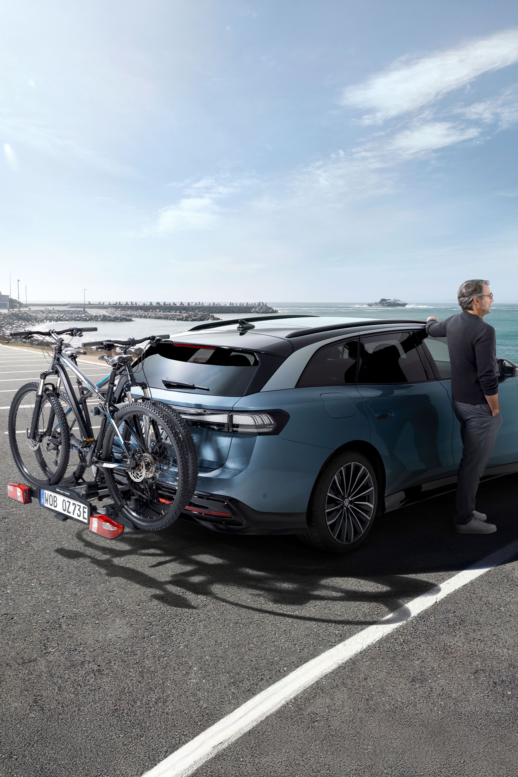 Muž stojí vedľa vozidla ID.7 Tourer s nosičom bicyklov VW a pozerá sa na more.
